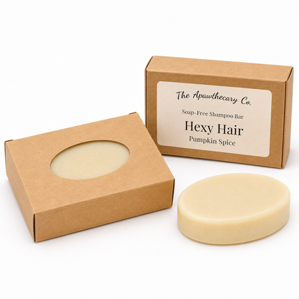 A round bar of soap-free shampoo bar in a cream color, shown next to its kraft paper packaging box with a circular window and another box displaying the label 'The Apothecary Co. Hexy Hair Pumpkin Spice'.