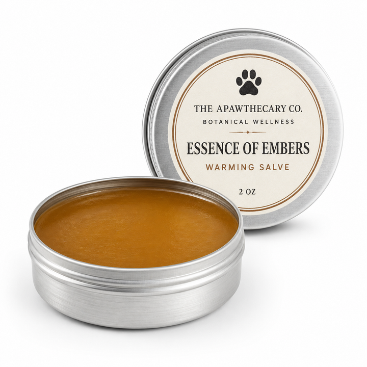 An open round silver tin of The Apawthecary Co. 'Essence of Embers' warming salve, showing the amber-brown balm inside, with its matching lid leaning behind it on a white background.