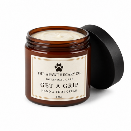An open amber glass jar of pet hand and foot cream with its black lid leaning against it, on a clean white background. The cream inside is off-white, and the label reads 'THE APAWTHECARY CO.
