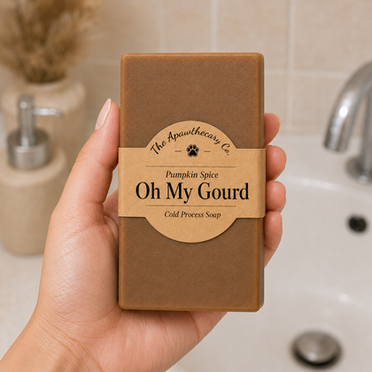 A hand holds a rectangular bar of soap in a bathroom setting. The soap is wrapped in brown paper with a tan paper label that reads 'The Apothecary Co.