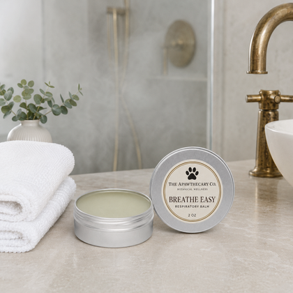 A silver round tin of Breathe Easy Respiratory Balm by The Apothecary Co. sits on a bathroom counter next to an open tin showing the balm inside, with a folded white towel, a small plant, and a brass faucet in the background.