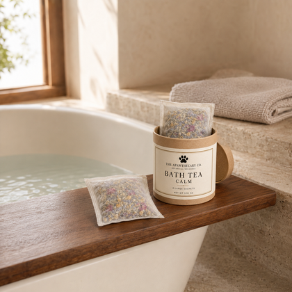 Bath tea calm in a kraft paper jar with a clear bag of loose herbal blend beside it, styled on a wooden bath ledge next to a bathtub in a warm, neutral bathroom setting.