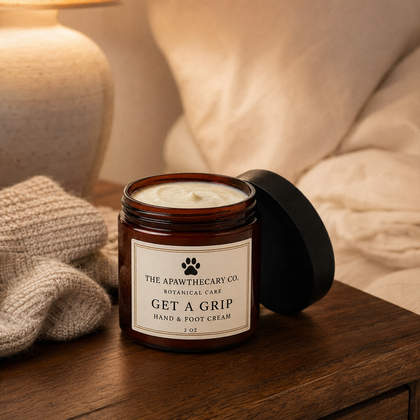 A candle in an amber glass jar with its black lid leaning against it, placed on a wooden surface in a cozy indoor setting with a knitted blanket and soft warm lighting in the background. The label reads 'THE APAWTHECARY CO.