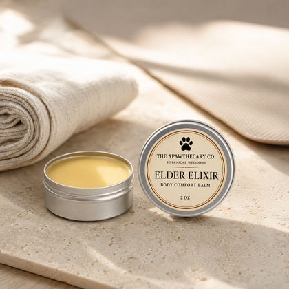 An open round silver tin of body comfort balm sits beside its matching lid on a stone surface, with rolled beige fabric in soft sunlight in the background. The lid reads 'THE APOTHECARY CO.
