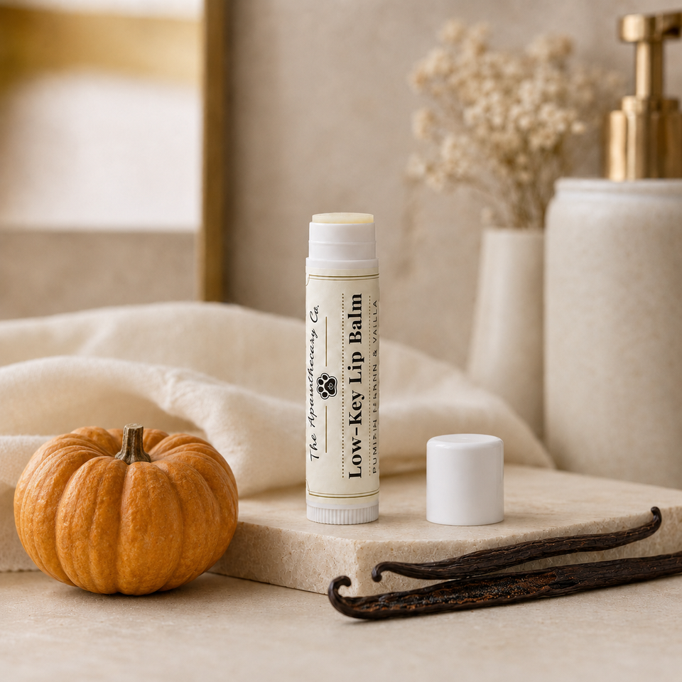 A tube of The Gentleman's Co. Low-Key Lip Balm stands on a stone slab beside its cap, with a small orange pumpkin, vanilla beans, and soft neutral-toned props in the background.