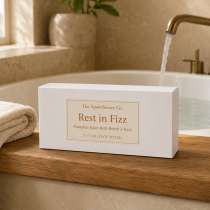 A white rectangular box of The Apawthecary Co. 'Rest in Fizz' pumpkin spice bath bomb 3-pack sits on a wooden ledge next to a folded towel, with a running faucet and bathtub in the background.