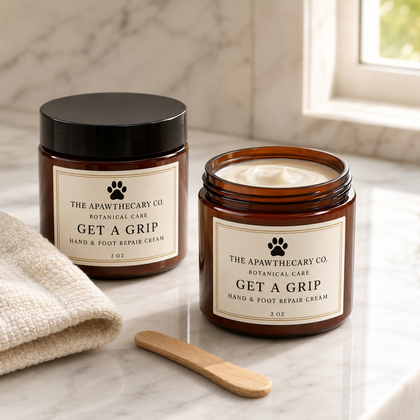 Two amber glass jars of The Pawthecary Co. Get A Grip hand & foot repair cream on a marble countertop, with one jar open to show the cream inside and a small wooden spatula in front.