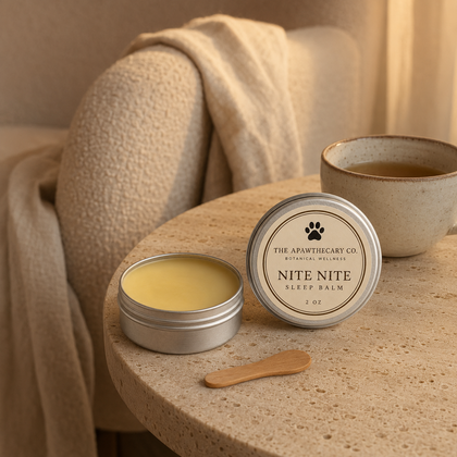 A silver round tin of Nite Nite sleep balm by The Apothecary Co. sits open on a speckled stone table, with its lid placed beside it and a small wooden spatula.