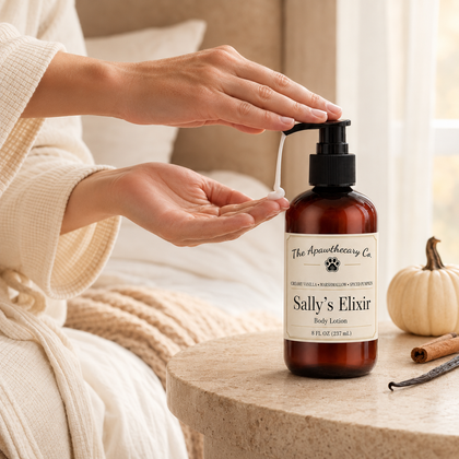 A person in a cream robe pumps body lotion from an amber bottle labeled 'The Apothecary Co. Sally's Elixir' onto their palm, with a small pumpkin and vanilla bean on a stone table nearby.