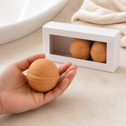 A hand holds a round brown bath bomb in the foreground, with a white box containing two more bath bombs behind it on a light beige surface.