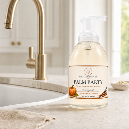 A bottle of Palm Party hand wash by The Apothecary Co. stands on a light stone countertop in a bright kitchen setting, with a gold faucet and sink in the background.