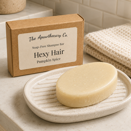 A bar of Hexy Hair soap-free shampoo by The Apawthecary Co. sits on a textured ceramic dish in front of its kraft paper packaging box, set on a bathroom counter with a folded towel and tiled wall in the background.