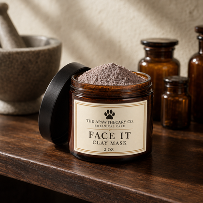 An open amber glass jar of The Apawthecary Co. Face It Clay Mask sits on a dark wooden table with its black lid leaning beside it.