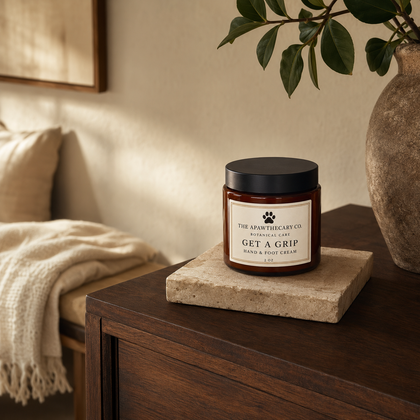 A jar candle labeled 'THE SPONTANEOUS DOG CO. GET A GRIP' sits on a stone coaster on a dark wooden table in a warm, softly lit room with a bed and plant in the background.
