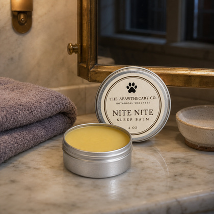 An open round silver tin of Nite Nite sleep balm sits on a marble bathroom counter beside a folded purple towel, with the lid placed behind it and a mirror in the background.
