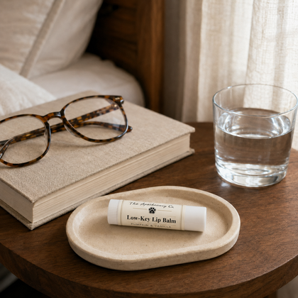 A tube of Low-Key Lip Balm by The Apothecary Co rests on a small beige dish on a wooden table, styled with a book, tortoiseshell glasses, and a glass of water in a cozy bedroom setting.