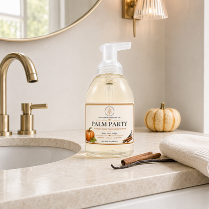 A bottle of The Artisan Soap Co. Palm Party hand wash soap sits on a beige marble bathroom counter next to a gold faucet, a rolled towel, and a small decorative pumpkin.