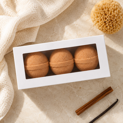 Three round brown bath bombs displayed in a white rectangular window box, styled on a beige stone surface with a cream towel, natural sponge, and vanilla bean.