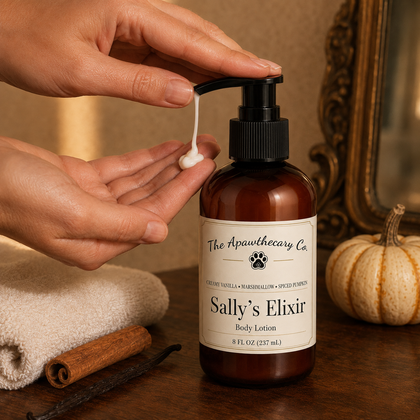 A person's hands are shown dispensing white body lotion from an amber pump bottle labeled 'Sally's Elixir' by 'The Apawthecary Co.' on a rustic wooden table with a cinnamon stick, vanilla bean, and a small pumpkin in the background.