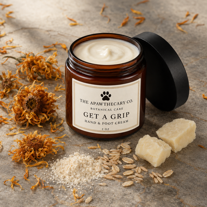 An amber glass jar of hand and foot cream with its black lid leaning against it, surrounded by dried orange flowers, oat flakes, and two cubes of cream on a textured stone surface. The label reads 'THE APAWTHECARY CO.