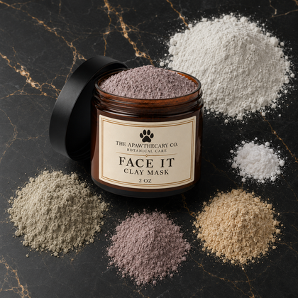 An open amber glass jar labeled 'FACE IT CLAY MASK' by The Apawthecary Co., surrounded by piles of different colored clay powder on a dark marble surface.
