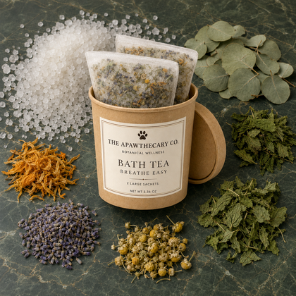 Two tea bags in a kraft paper container labeled 'The Apawthecary Botanical Wellness Bath Tea' on a dark green stone surface, surrounded by loose botanical ingredients including eucalyptus leaves, dried herbs, lavender, chamomile, and sea salt.