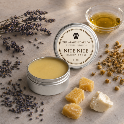 An open round silver tin of Nite Nite Sleep Balm by The Apawthecary Co. is shown on a neutral stone surface with lavender sprigs, dried flowers, honeycomb pieces, and a small glass bowl of golden oil.