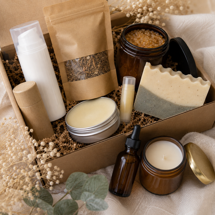 A curated skincare gift set arranged in a kraft box with decorative dried flowers and greenery. The set includes a white pump bottle, a brown stand-up pouch with a clear window showing loose botanicals, an amber bath bomb jar, a layered soap bar, a silver tin of cream, a lip balm tube, a dropper bottle, and a candle jar with a gold lid.