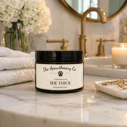 Jar of 'She Thick' whipped body butter from The Apawtheery Co on a bathroom counter with flowers and candles in the background.