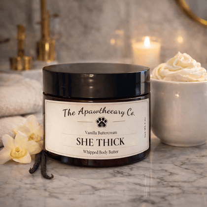 Jar of 'She Thick' whipped body butter from The Apawtheery Co. on a marble surface with vanilla flowers and a candle in the background.