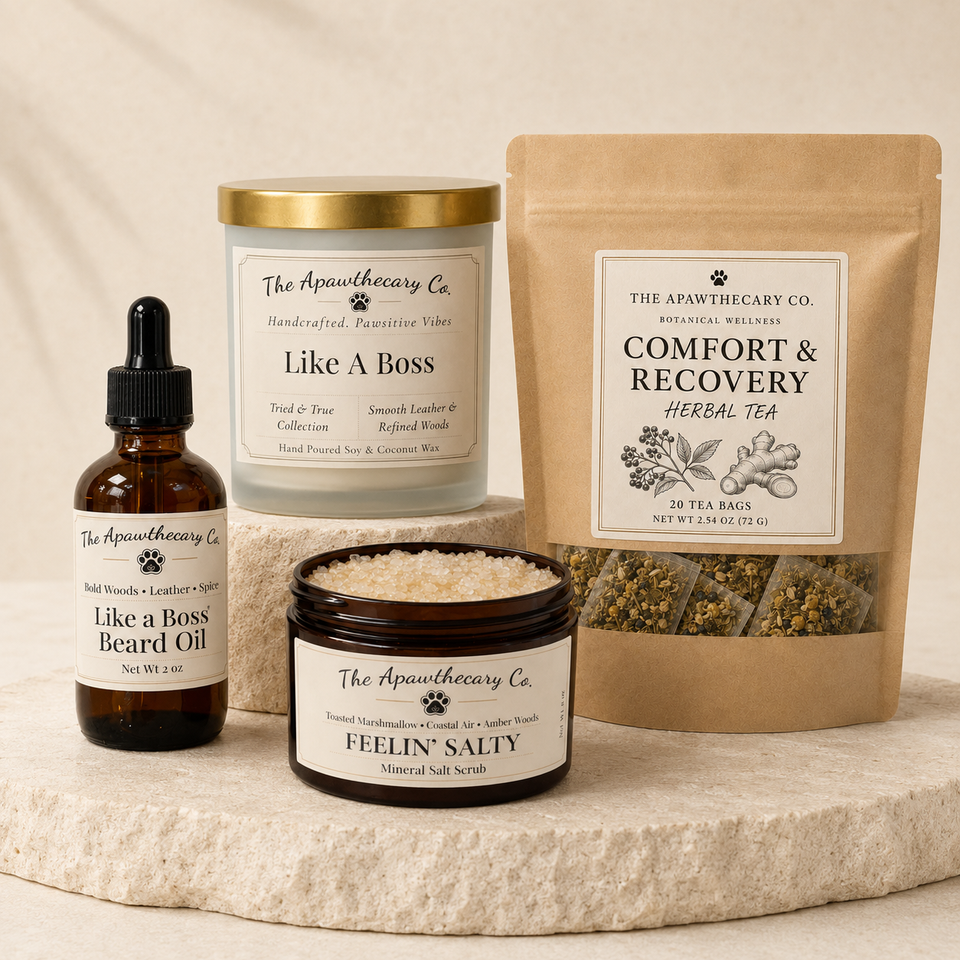 Four skincare and wellness products from The Apathhecy Co. arranged on a light stone surface against a soft beige background: a jar of Like A Boss handcrafted body wash, a pouch of Comfort & Recovery Herbal Tea, a bottle of Like a Boss Beard Oil, and a jar of Feelin' Salty mineral salt scrubs.
