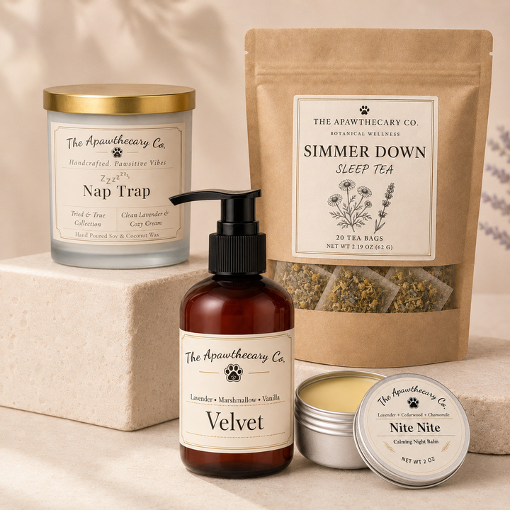 A curated product set from The Apawthecary Co. arranged on a light beige surface, including a candle in a glass jar with a gold lid, a brown pump bottle labeled Velvet, a kraft stand-up pouch of Simmer Down Sleep Tea, a small round tin labeled Nite Nite, and a cream block behind the candle.