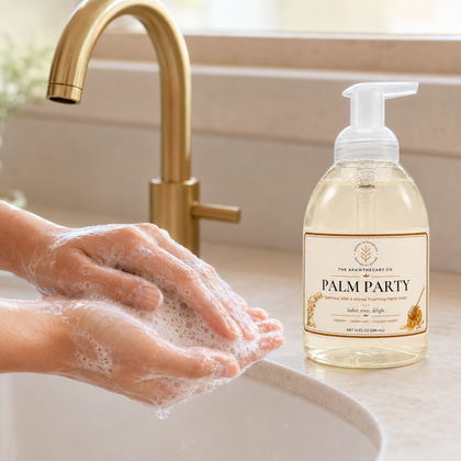 A bottle of Palm Party hand soap by The Art of Soap Co. sits on a bathroom counter next to a sink with a gold faucet, while a person's hands are shown lathering soap foam.