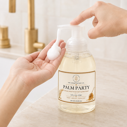 A clear pump bottle of palm soap with a white label reading 'The Artisanal Co. Palm Party' is shown on a bathroom counter, with a hand pressing the pump and another hand holding a dollop of the white foam.