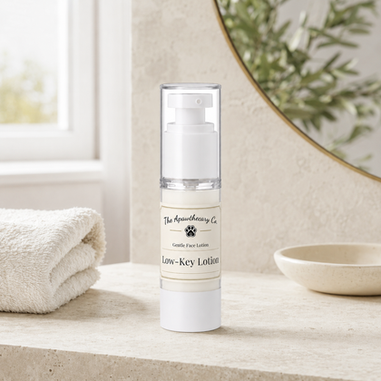 A clear pump bottle of Low-Key Lotion by The Apawthecary Co. stands on a stone surface in a bathroom setting, with a folded towel, a small dish, and a mirror in the background.
