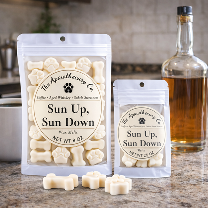 Two packages of 'Sun Up, Sun Down' wax melts from The Apawthecary Co. on a kitchen counter with a bottle of whiskey in the background.