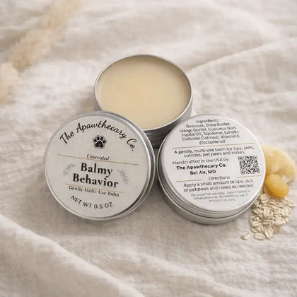 Two tins of 'Balmy Behavior' balm from The Apawthecary Co. on a soft fabric background.