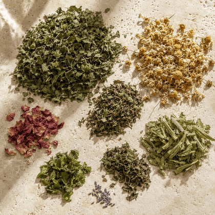 A flat-lay arrangement of various dried and fresh botanical ingredients, including green herbs, yellow chamomile flowers, pink rose petals, and purple lavender sprigs, displayed on a textured beige stone surface.