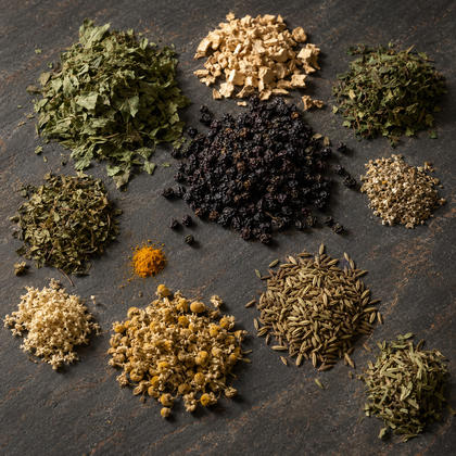 An overhead flat-lay arrangement of various loose-leaf herbal teas and spices on a dark textured slate background, including green tea, chamomile, black seed, cumin, turmeric, and dried roots.