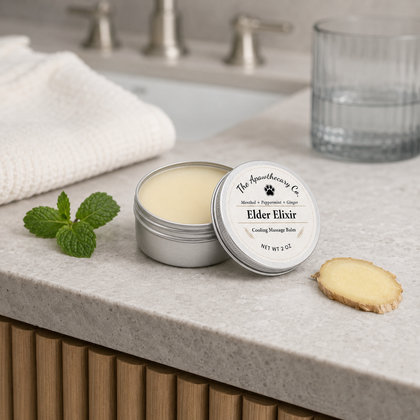 Open tin of Elder Elixir balm beside mint, ginger, a folded towel, and a glass of water.