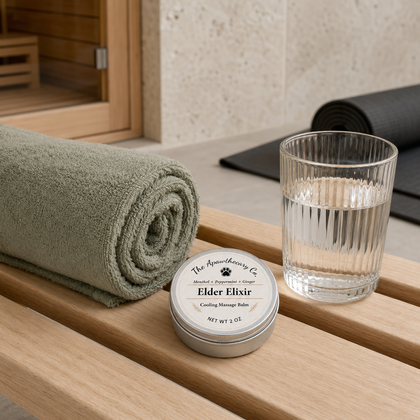 Elder Elixir balm tin beside a rolled green towel and a glass of water on a wooden bench.