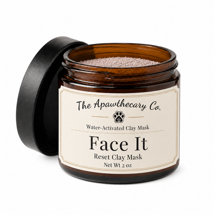 An open amber glass jar of The Apawthecary Co. Face It Water-Activated Clay Mask, showing the light brown clay powder inside, with its black lid leaning against it on a white background.