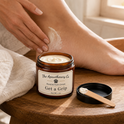 A person applies white hand and foot cream from an amber glass jar labeled 'The Apawthecary Co. Get a Grip' while sitting at a wooden table, with the jar lid and a small wooden spatula beside it.