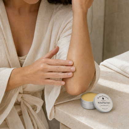 A woman in a cream linen robe applies herbal skin salve from an open silver tin to her forearm, with the product tin resting on a stone counter nearby.