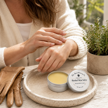 A person applies a yellow herbal skin salve from a small silver tin resting on a round stone tray. The scene includes brown leather gloves, a potted green plant, and the person wearing a cream cardigan.