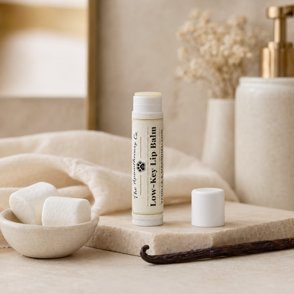 Low-Key Lip Balm tube beside its cap, marshmallows, and a vanilla bean on a stone surface.