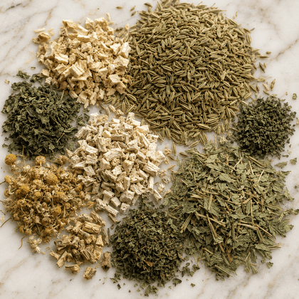 An assortment of dried and chopped herbs and spices arranged in small piles on a white marble surface. The mix includes green leafy herbs, a pile of light beige dried roots or bark, yellow dried flowers, and brown seeds or grains.