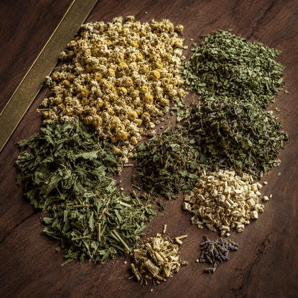 Various loose-leaf herbal teas and botanical blends arranged in circular piles on a dark wooden surface. The blends include yellow and white dried flowers, green leafy herbs, dried green herbs, light beige root pieces, and small purple flower buds.