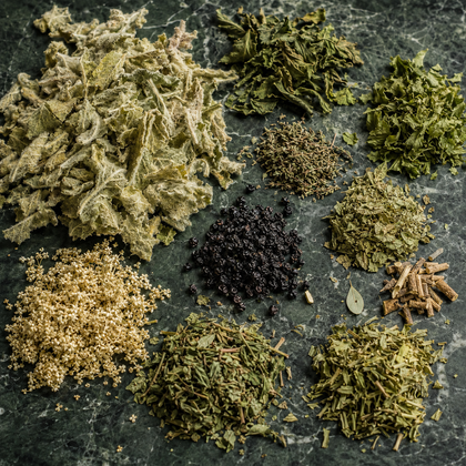 An assortment of dried and ground herbs and spices arranged in small piles on a dark green marble surface. The collection includes various green leafy herbs, a pile of pale beige seeds or grains, a central dark brown/black berry-like spice, and a small piece of dried bay leaf.
