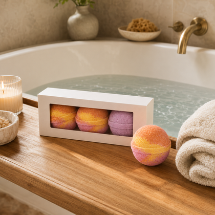 A boxed set of three bath bombs with one loose bath bomb in front, displayed on a wooden bathtub ledge with a rolled towel, candle, and bathwater in the background.